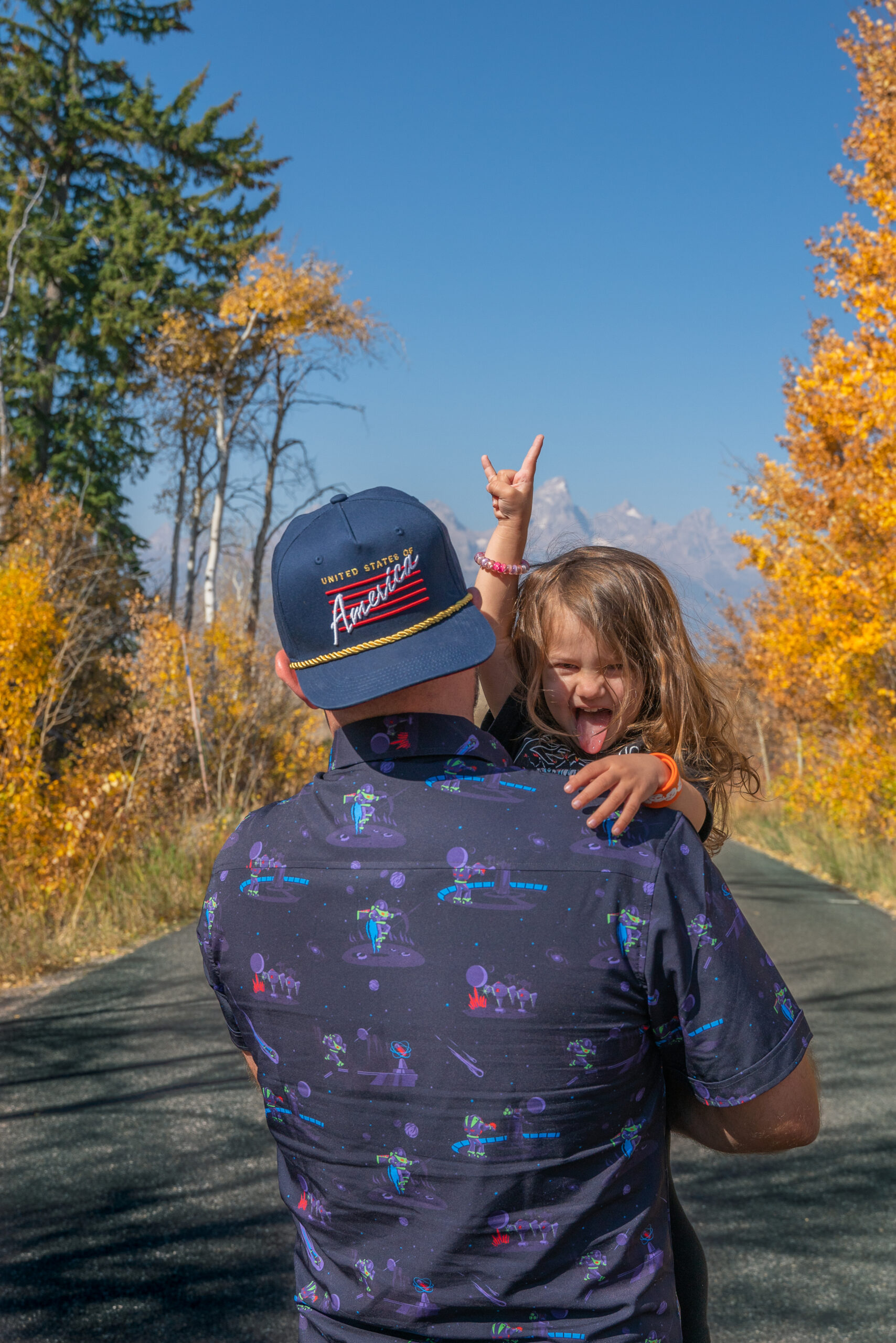 Child holds up a rock and roll sign with tongue out over father's shoulder as he walks through fall in Grand Tetons National Park